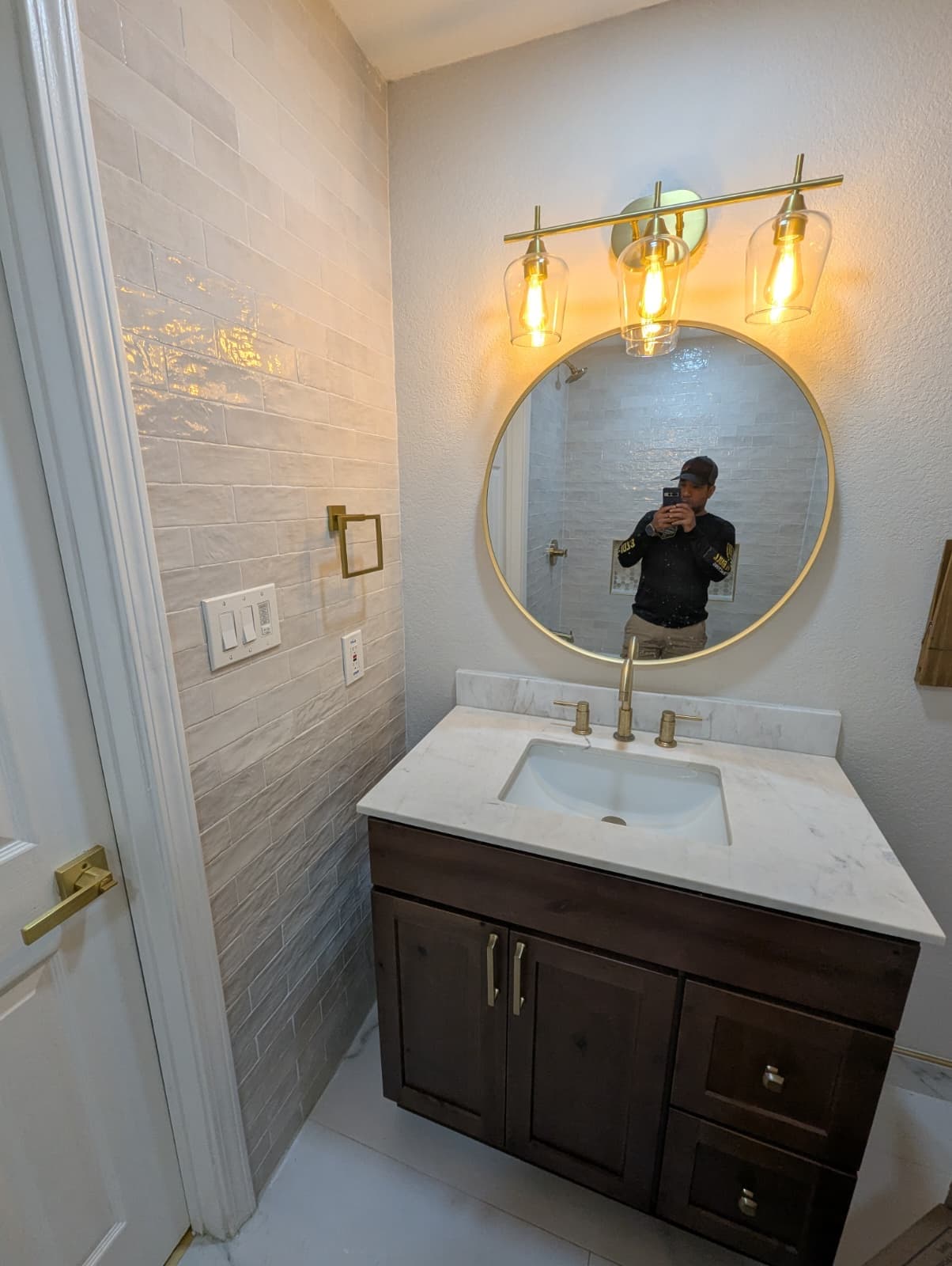 Bathroom remodel in Alamo Ranch San Antonio TX by Raging Bull Contracting — dark wood vanity with marble-look quartz top, round brass mirror and three-light brass fixture against a glossy zellige-style tile wall