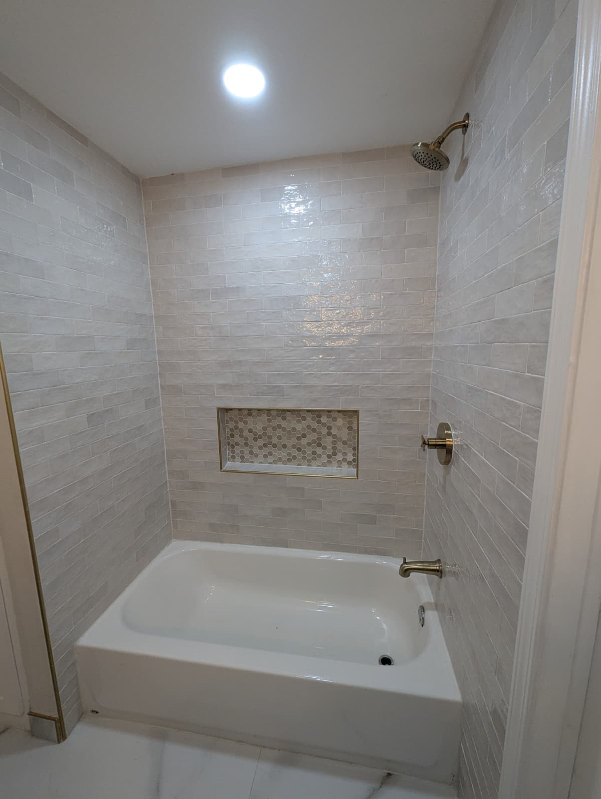 Bathroom remodel in Alamo Ranch San Antonio TX by Raging Bull Contracting — white alcove tub under glossy cream zellige-style subway tile with a brass-trimmed hex mosaic niche and brass shower fixtures