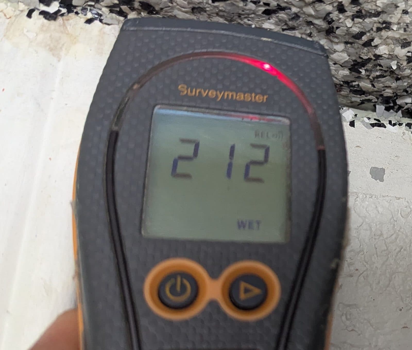 Moisture meter reading 212 REL with the WET indicator still lit at the garage floor and wall joint, next room over