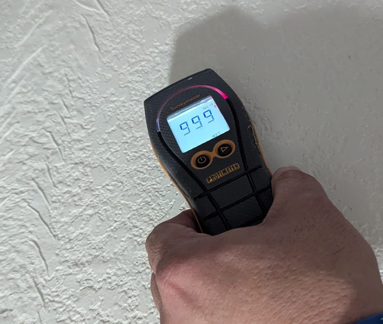 Moisture meter reading 999 REL with the WET indicator lit, held against a laundry-room wall in Cibolo TX